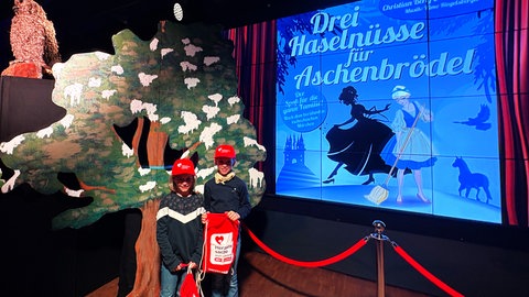 Zwei Kinder mit Herzenssache-Turnbeuteln und -Mützen stehen neben vor dem Musical-Plakat