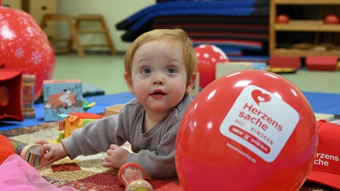 Clara liegt auf einer Spielmatte, vor ihr ein roter Herzenssache-Luftballon