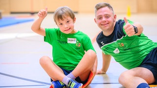 Timo und Valentin sitzen in der Sporthalle und zeigen mit dem Daumen nach oben
